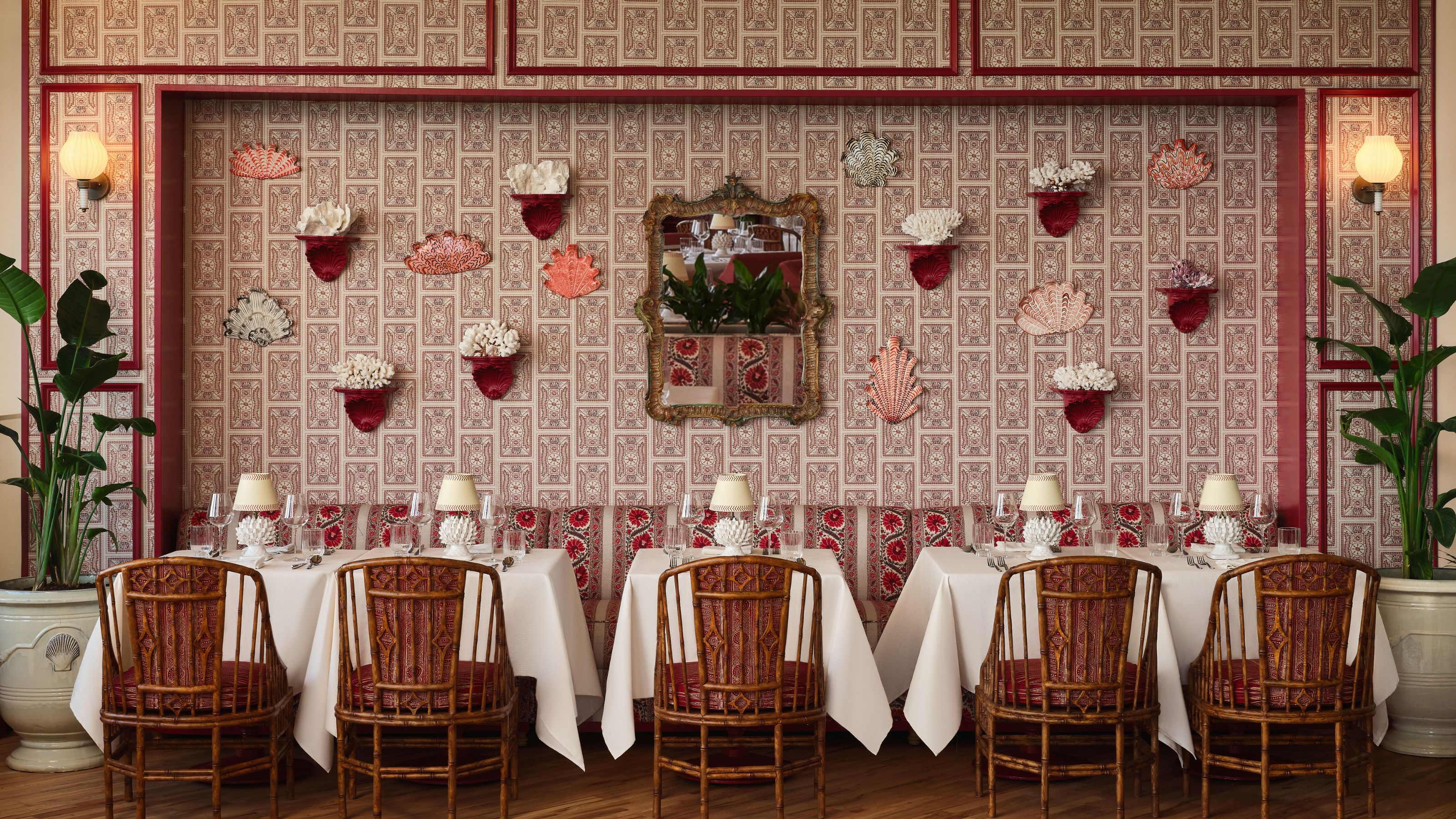 A dining room setup featuring three tables with white tablecloths and high-backed bamboo chairs, set against a red patterned wall decorated with an ornate mirror and coral sculptures on small shelves.
