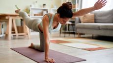 woman performing a bird dog exercise from her hands and knees on an exercise mat in a living room setting. she's looking down at the floor facing the camera.