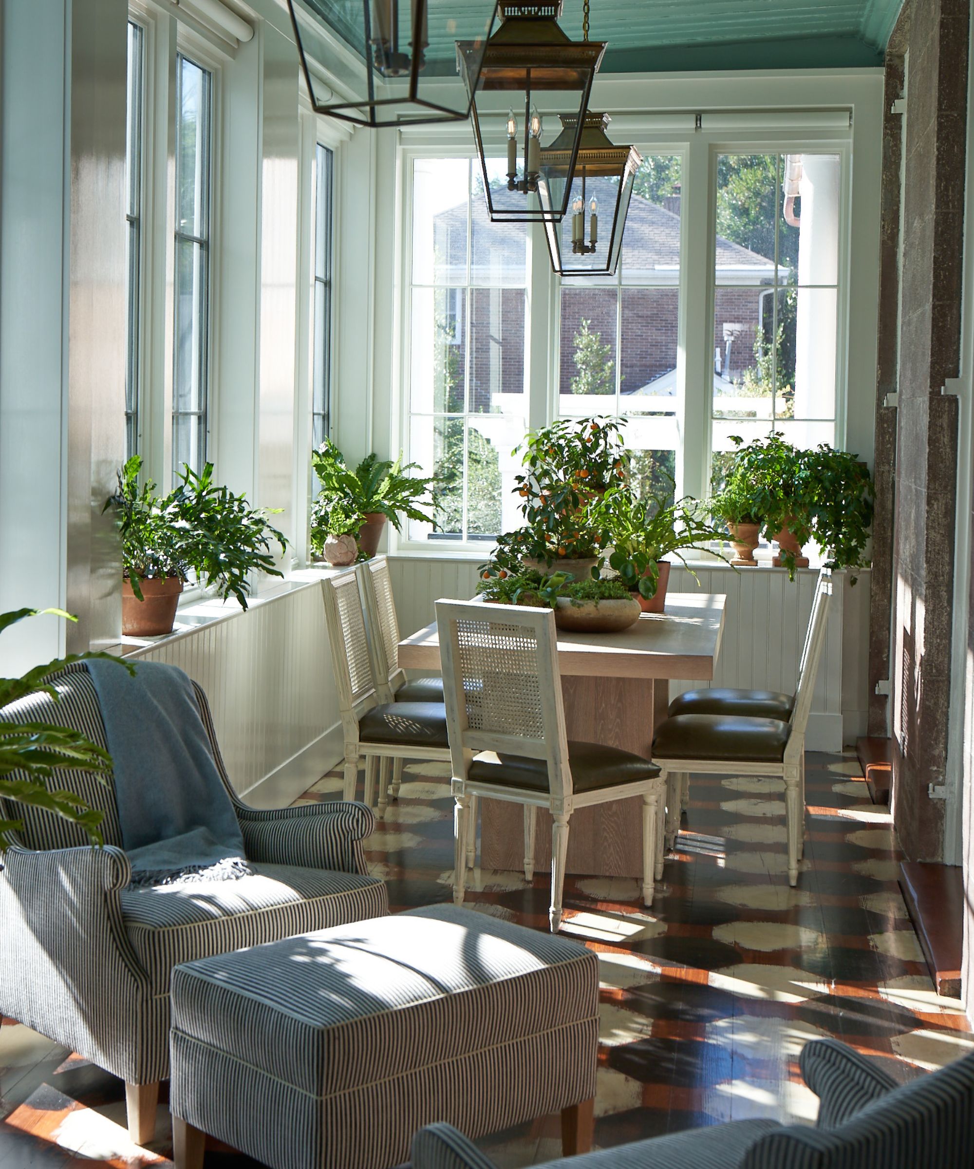 Conservatory with tiled floor, glass windows, three glass lantern lights on the ceiling, a dining table with potted plants on top, dining chairs, and a striped armchair and stool in the corner.