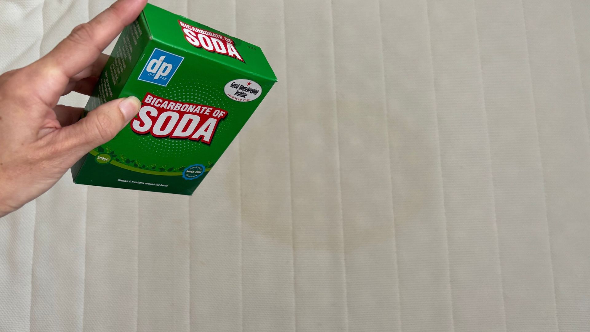 The image shows a hand holding a green baking soda box, which is being sprinkled on top of a yellow mattress stain