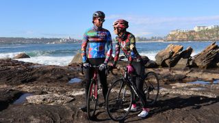 A pair of cyclists stand on the rocks on a beach, wearing loud-patterned cycology jerseys