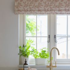 a kitchen with white walls and a fluted front Belfast sink and gold hardware with a pink and cream patterned roman blind