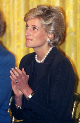 Princess Diana sitting in a chair against a gold curtain clapping and wearing a black dress with pearls and gold Cartier watch