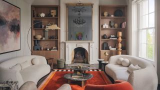 Living room with pale grey walls, white curved twin sofas, bright orange rug, black marble coffee table and wooden built-in shelving either side of a fireplace