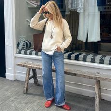 British style influencer Chloe Butler stands outside a shop in London wearing a cream sweater, pendant cord necklace, suede tote bag, cuffed jeans, and red jelly flats