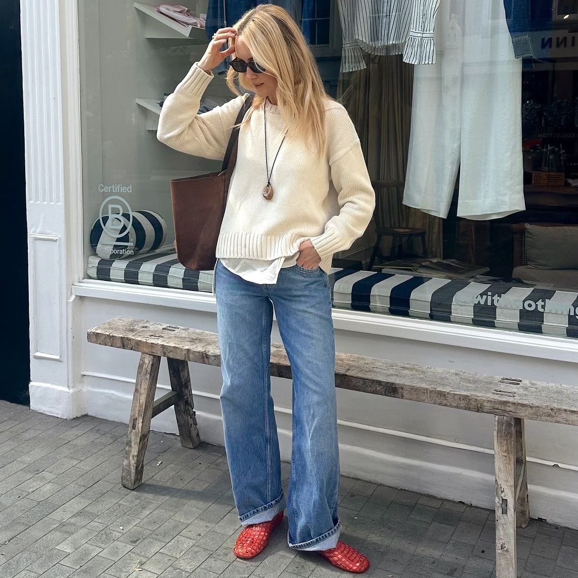 British style influencer Chloe Butler stands outside a shop in London wearing a cream sweater, pendant cord necklace, suede tote bag, cuffed jeans, and red jelly flats