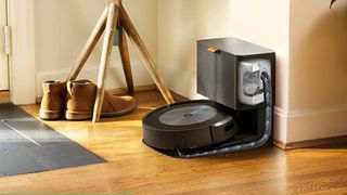 The Roomba j7+ in its bases station in front of a table with shoes underneath the table's legs