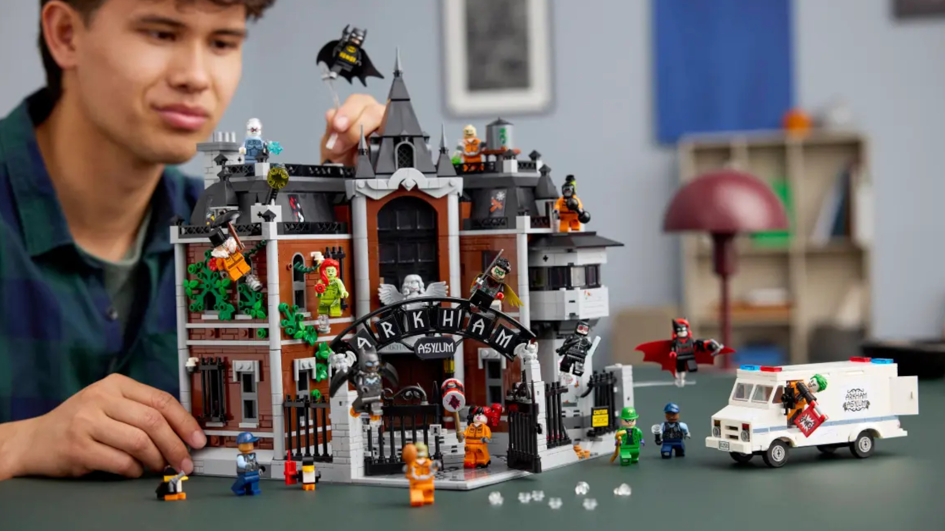 A man smiles as he places a Batman minifigure on a plain stand at the top of the Lego Arkham Asylum set, which is sat on a dark surface