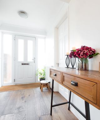 A bright entryway with white walls, windows and a white door, and a brown console table with purple flowers on it