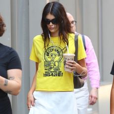 Kaia Gerber wears a yellow t-shirt with a white skirt and classic ballet flats while walking in new york city