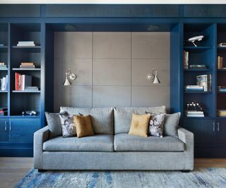 blue built in bookshelves with inset space for grey sofa, a grey tiled wall and wall lights