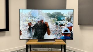 65-inch LG C4 TV photographed straight-on on a wooden stand. On the screen is an image of a golfer being sprayed with champagne.