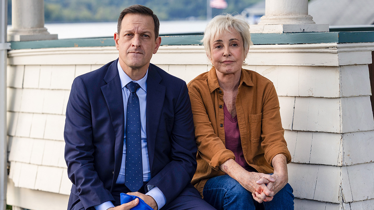 Still of Josh Charles sitting on a porch in a suit next to co-star Annie Potts in a brown jacket.