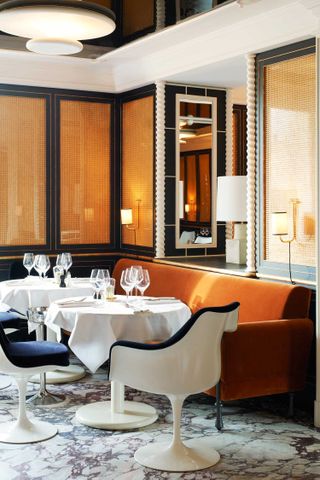 A modernly furnished restaurant features marbled floors, futuristic white seating and orange banquettes, rattan walls, and warm lighting.