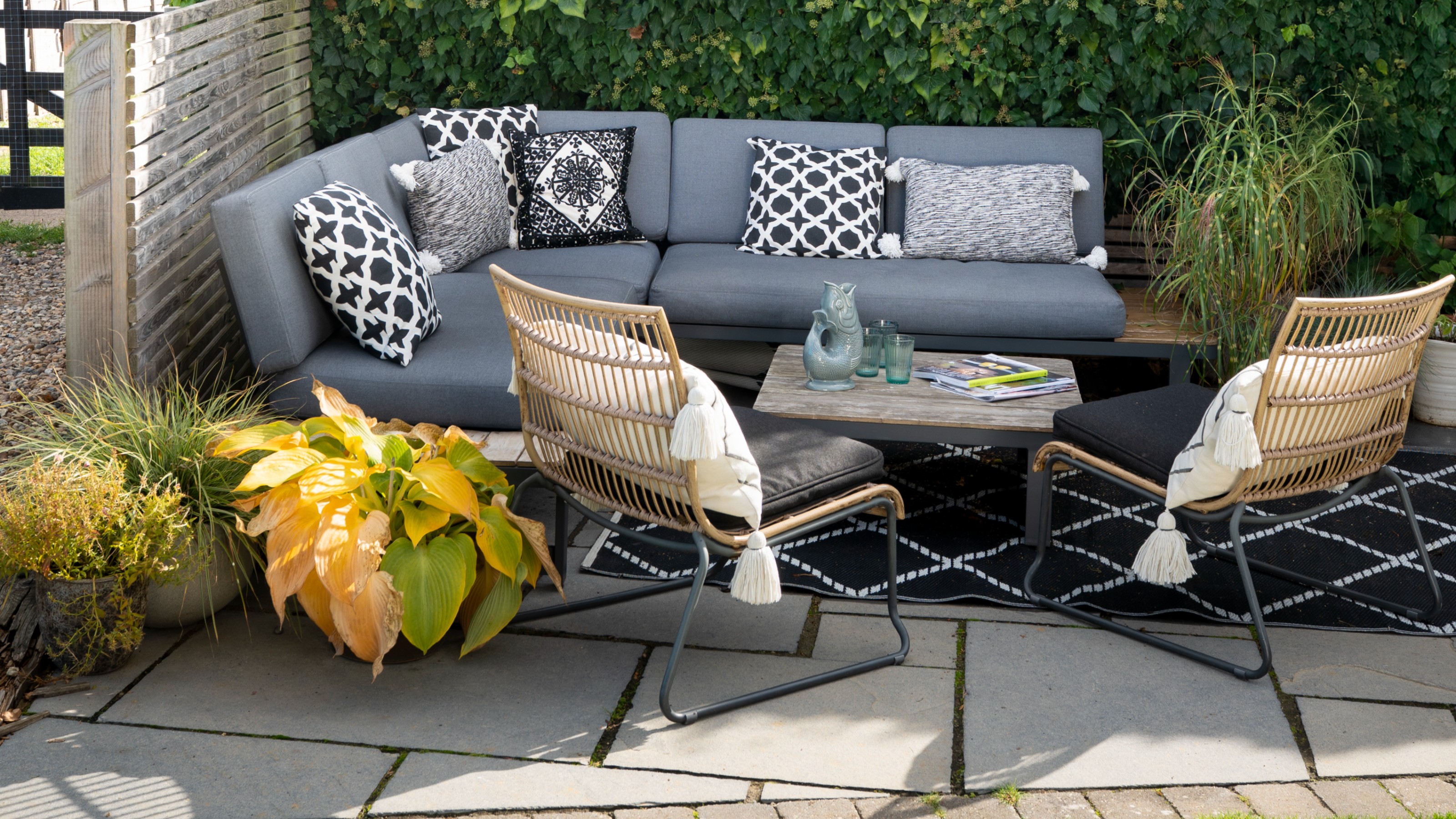 Agarden with a corner outdoor sofa with grey cushions and patterned scatter cushions and two rattan garden chairs