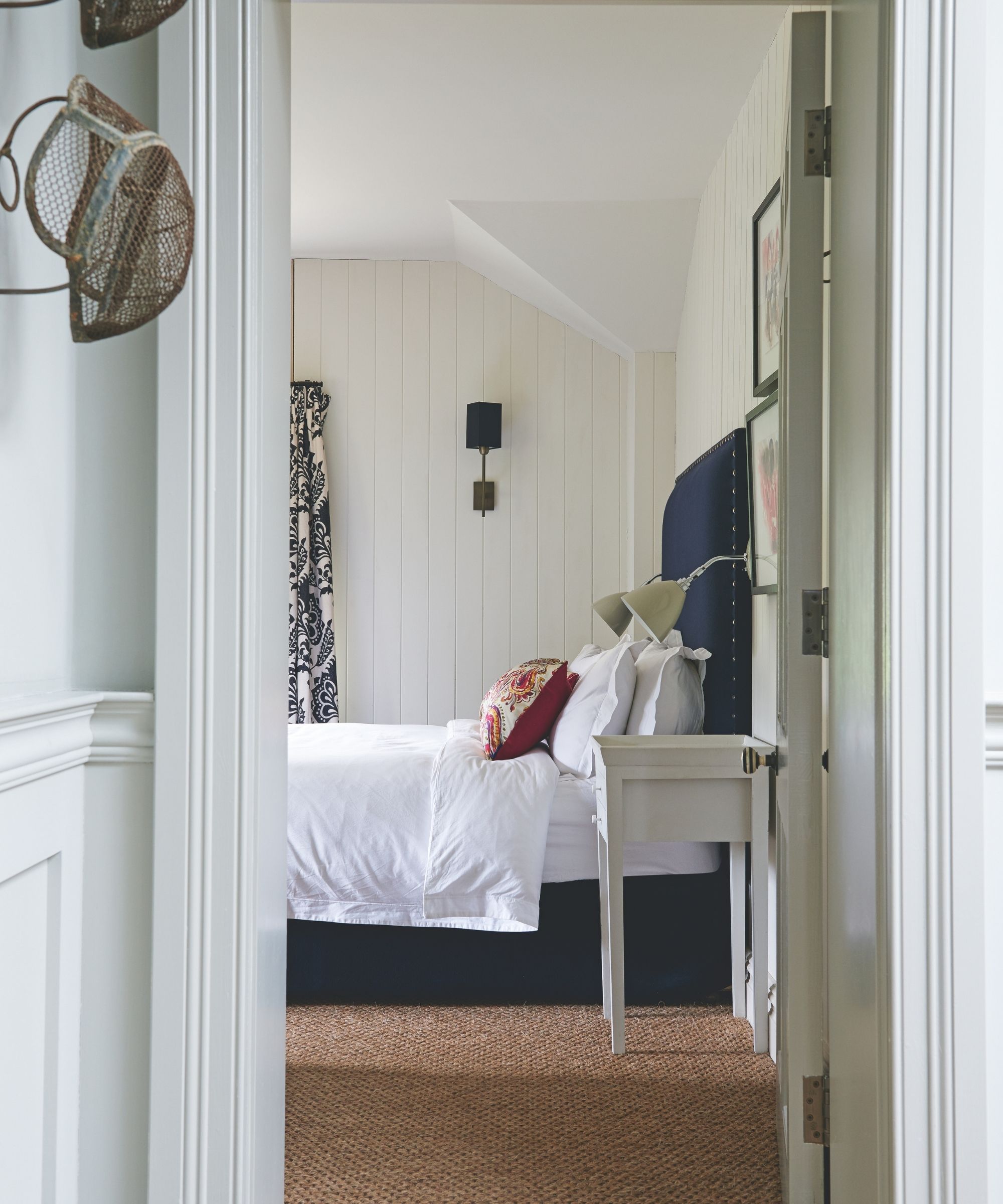 White panelled bedroom seen through an open doorway with a dark blue bed with white bedding, jute carpet, gallery wall and black and white patterned curtains.