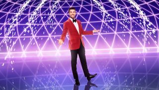 Joel Dommett stands in front of a purple background with pyrotechnics erupting behind him, wearing a red velvet tuxedo jacket and black trousers