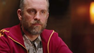 John Malkovich wearing a red track jacket looking serious.