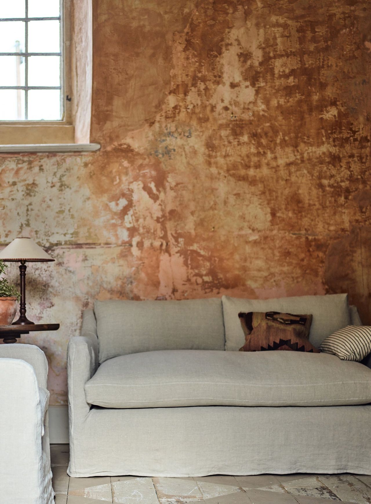 Warren Loose Cover Sofa in Flax Linen on a sofa in a room with a rustic, copper-colored wall behind it.