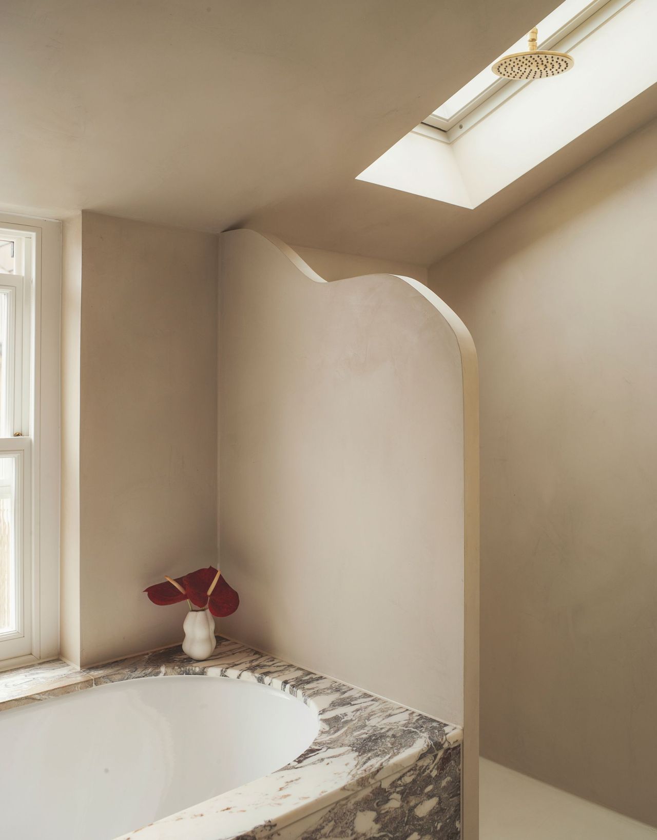A modern neutral bathroom with a marble bathtub and curved wall between the shower with a vase with a red flower at the side of the bath