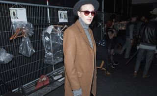 Man wearing a brown long coat, sunglasses and hat posing for the camera