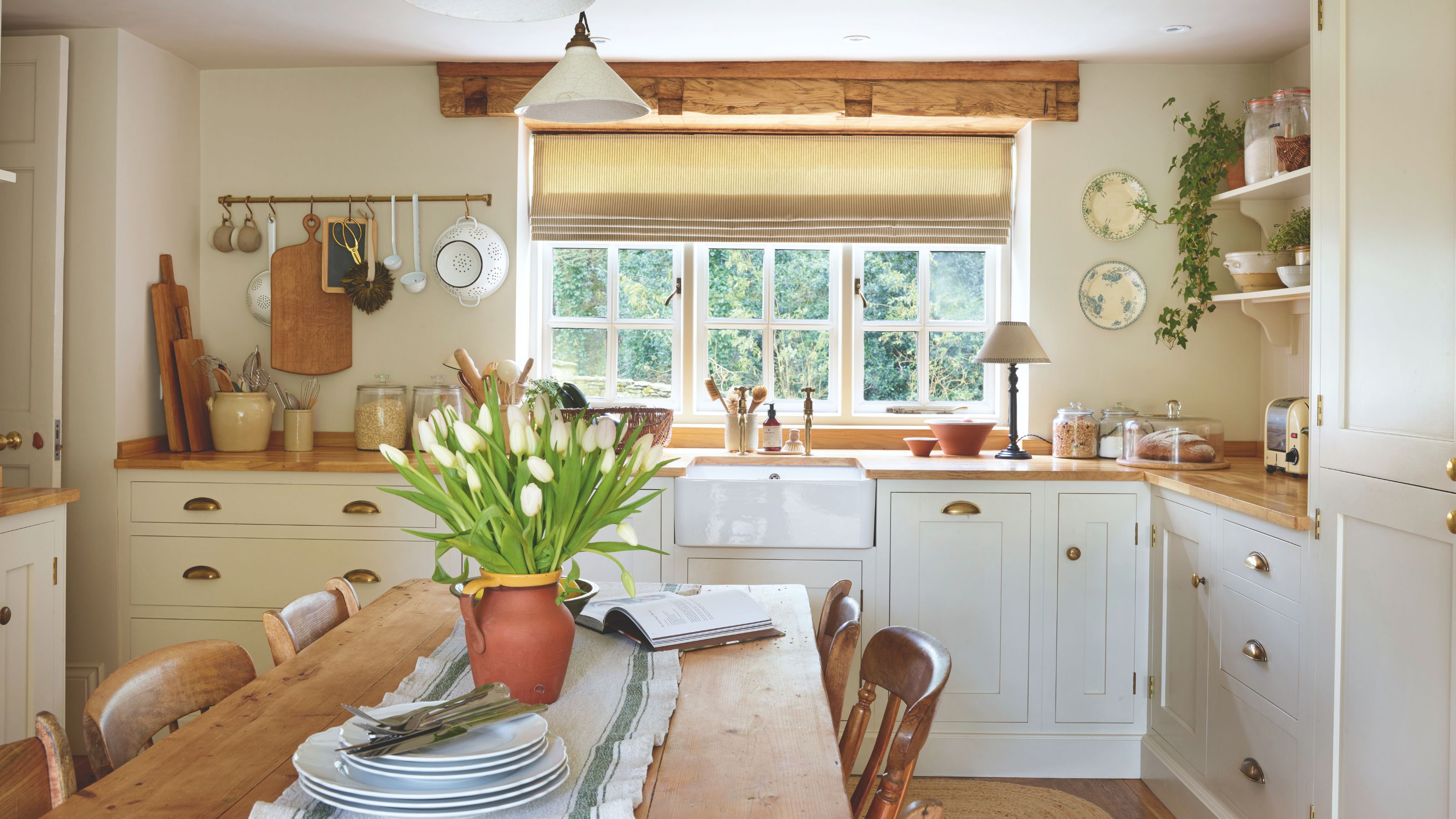 A country kitchen with cream cabinets and wooden worktops. A terracotta vase of white tulips on a wooden kitchen table in the centre of the room, and a white farmhouse sink beneath a window behind it.