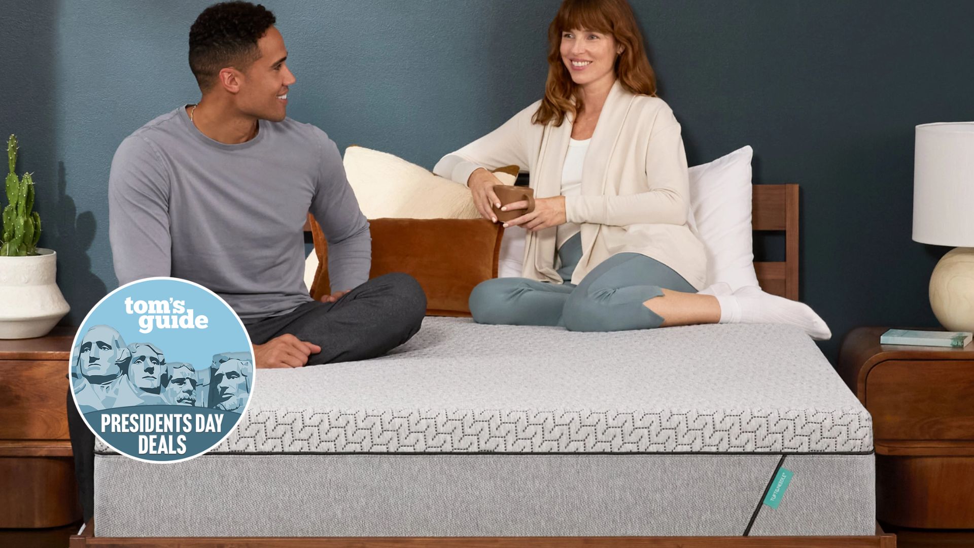The image shows two mattress testers sat on top of a grey mattress, placed on a wooden bed frame. Overlaid on top is a Tom's Guide Presidents' Day Deals badge