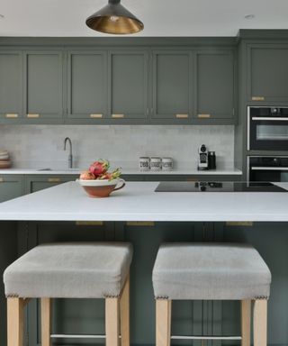A kitchen with concrete gray cabinets with gold handles, an island with a white countertop and gray panels, and two gray fabric stools with light wooden legs