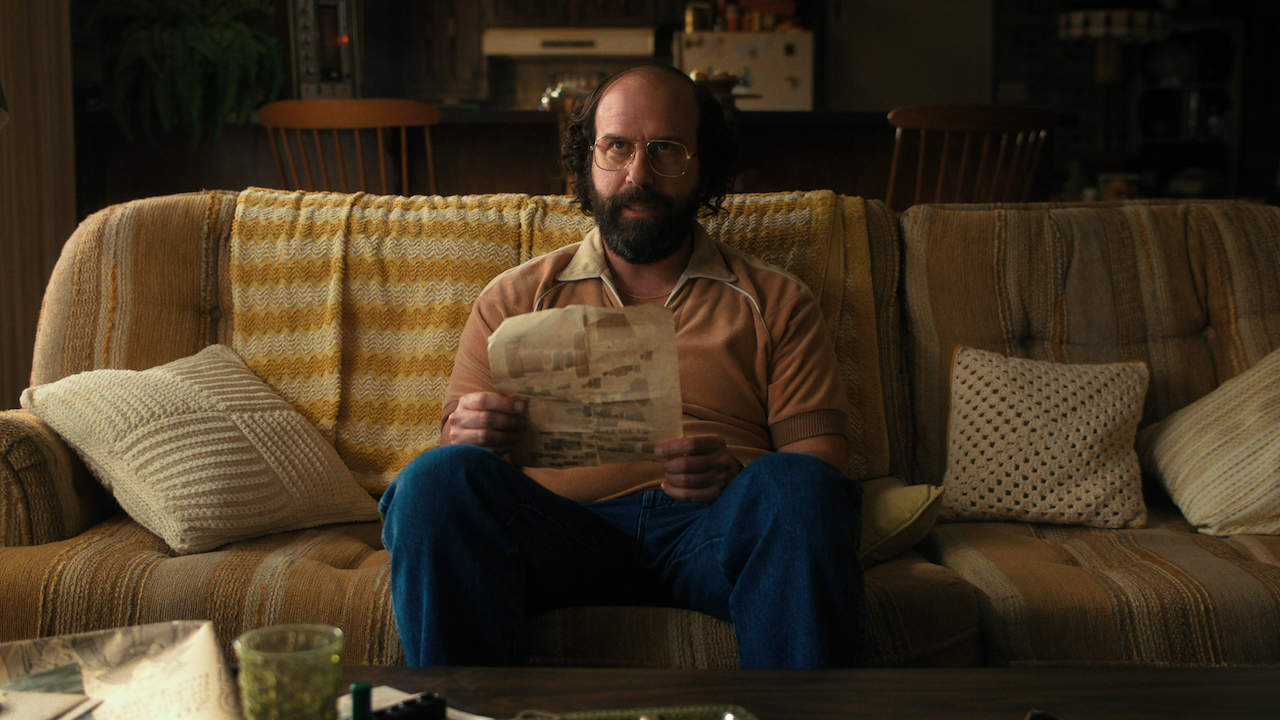 Brett Gelman&#039;s Murray Bauman seated on couch and looking at a piece of paper in his hands in Stranger Things