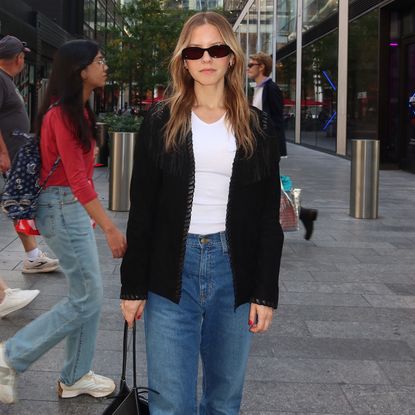 Nikki wears a white top, black jacket, and jeans.