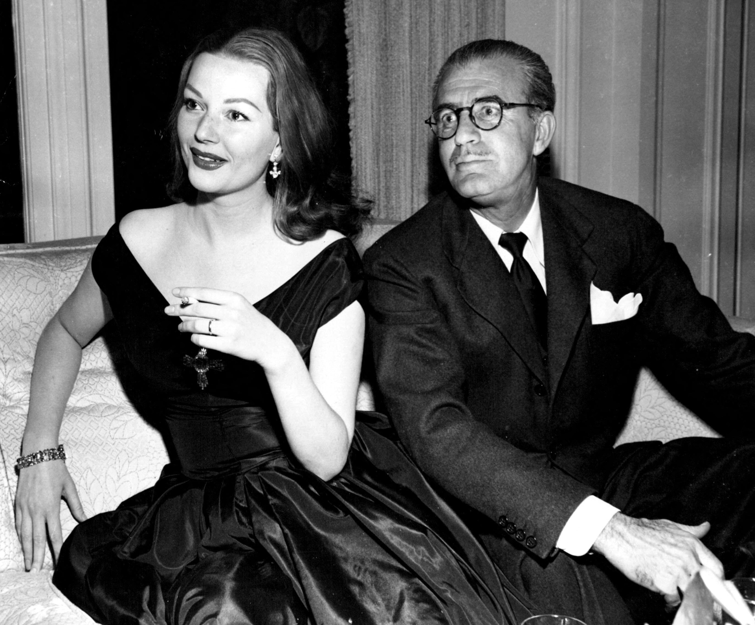 A black and white photograph of a woman and a man, sitting on a sofa, dressed in cocktail attire