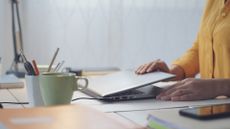 A business person closing their laptop, which sits on a cluttered desk.