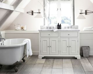 Bathroom with gray wooden flooring, gray cabinet with twin sinks positioned below window, twin wall lights either side of window, sloped ceiling with white bathtub to the left