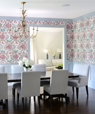 Colorful dining room with floral wallpaper, sky blue painted paneling and trim, gold pineapple pendant light, dark wood flooring and rectangular dining table, gray upholstered dining chairs