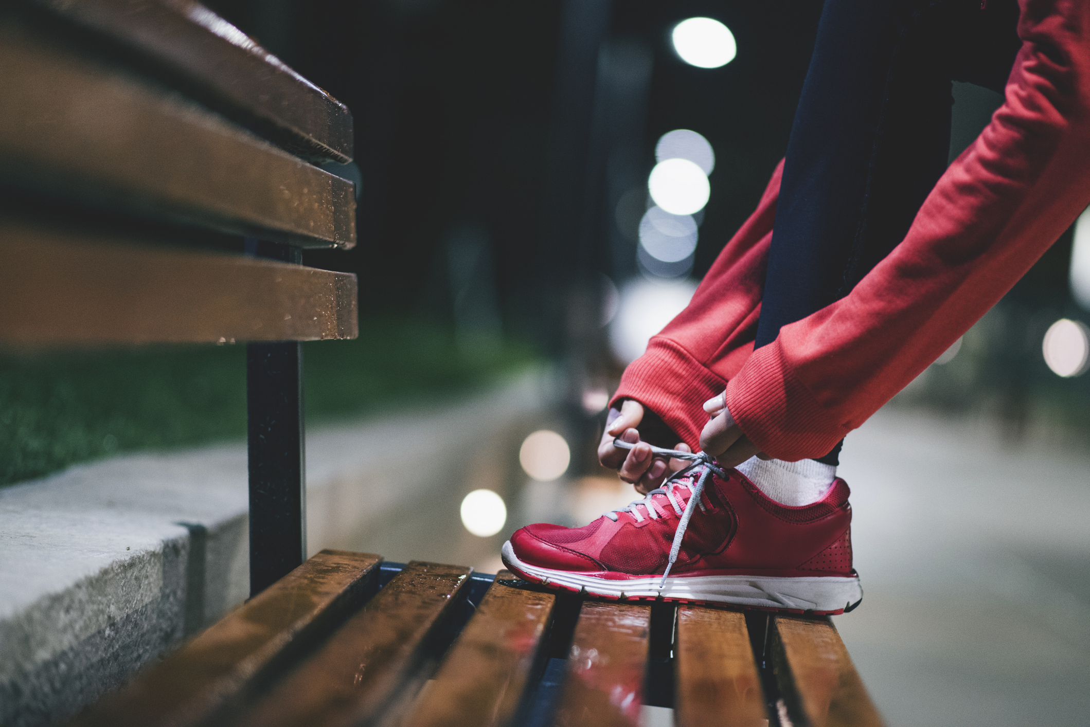 A person tying the laces of their running shoes while their foot rests on a wooden bench at night.