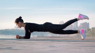 Woman performing a forearm plank outdoors by the ocean with left leg lifted in the air
