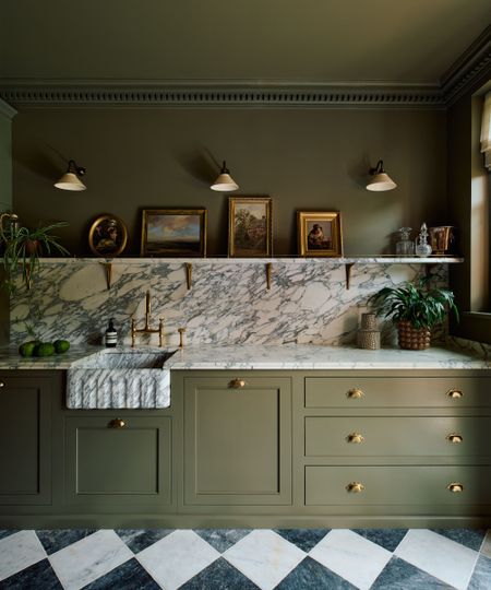 A classic kitchen with olive green cabinets and matching wall and ceiling, marble countertops and backsplash, a gallery shelf with antique picture frames, and sconce lighting on the wall. 