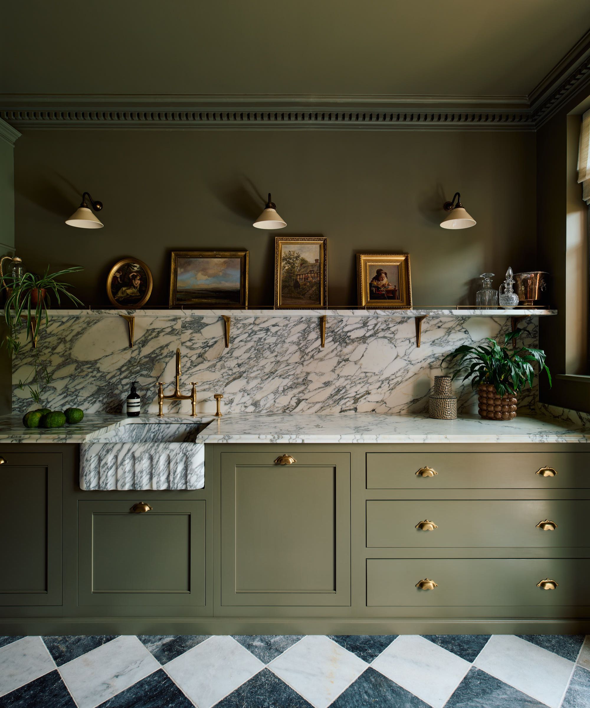 A classic kitchen with olive green cabinets and matching wall and ceiling, marble countertops and backsplash, a gallery shelf with antique picture frames, and sconce lighting on the wall.