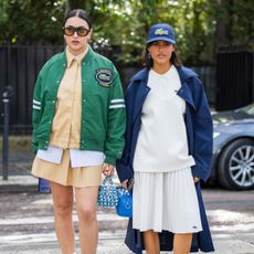 A guest wears green college jacket, beige button shirt, skirt & a guest wears cap, blue coat, white pleated skirt, shirt outside Lacoste during the Womenswear Fall/Winter 2024/2025 as part of Paris Fashion Week on March 05, 2024 in Paris, France.