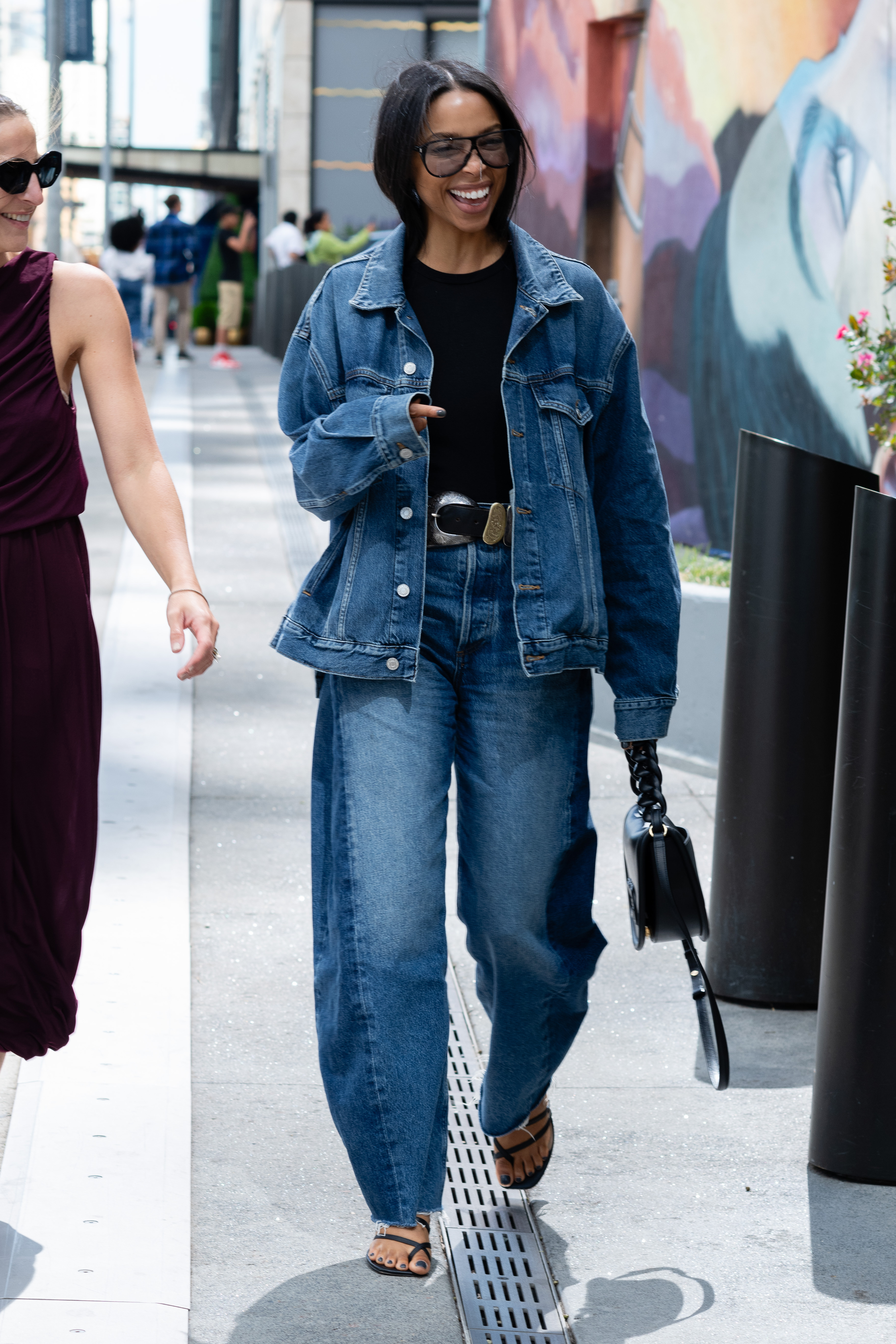 A woman is seen wearing a blue denim jacket, blue denim pant, black shirt, black sandals, black and silver belt, black sunglasses and carrying a black bag during New York Fashion Week on September 08, 2024.