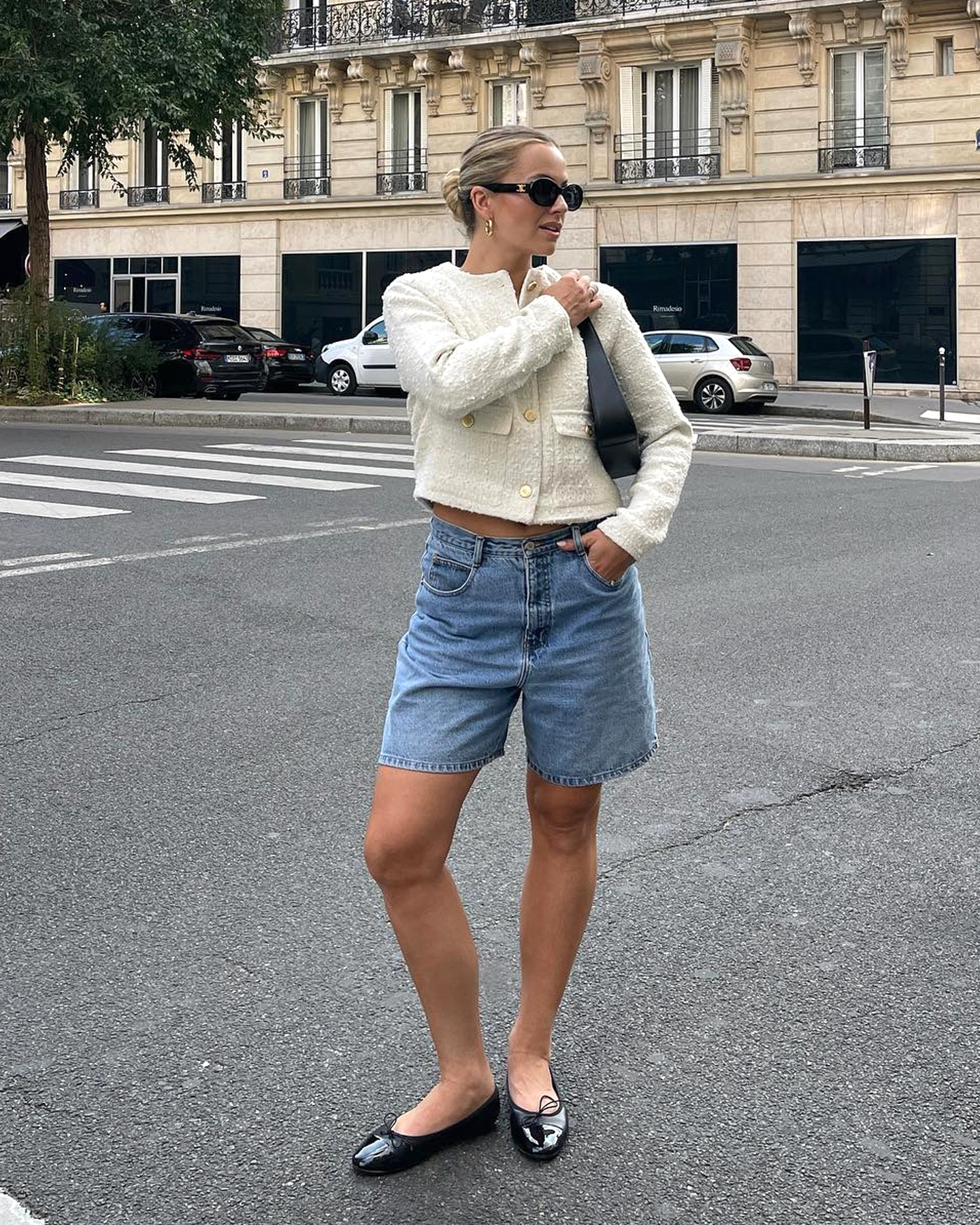 Style influencer Izzy Dilger poses on a street in Paris wearing black oval sunglasses, a cream tweed jacket, black mini shoulder bag, long denim shorts, and black patent ballet flats.