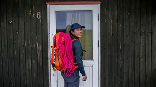 A woman stands in front of a wooden building, turning to look at the camera. She is wearing a bright orange Salewa Ortles Wall 38L Backpack with bright pink ropes attached.