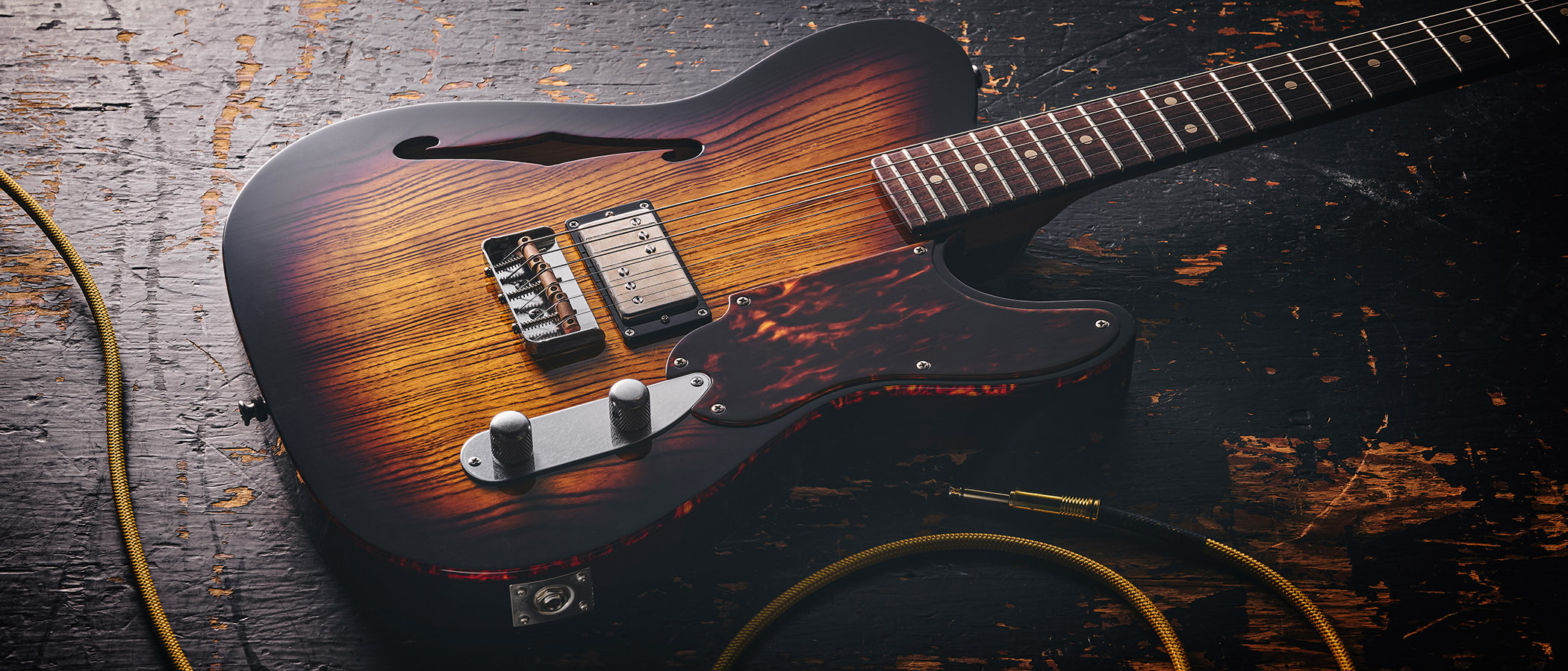 Patrick James Eggle Oz-t Cabronita Thinline; this semi-hollow T-style has a dark sunburst that reveals all the open grain, complementing the tortoiseshell pickguard, and is photographed here against a distressed floor, white and wooden backgrounds.