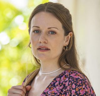 Cara Theobold in character as Rose Dalton, wearing a dark blue dress with a pink and orange floral print, and with a tote bang slung over her shoulder