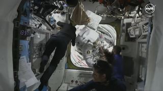 an astronaut in a bulky white spacesuit is brought into a cramped laboratory from an airlock