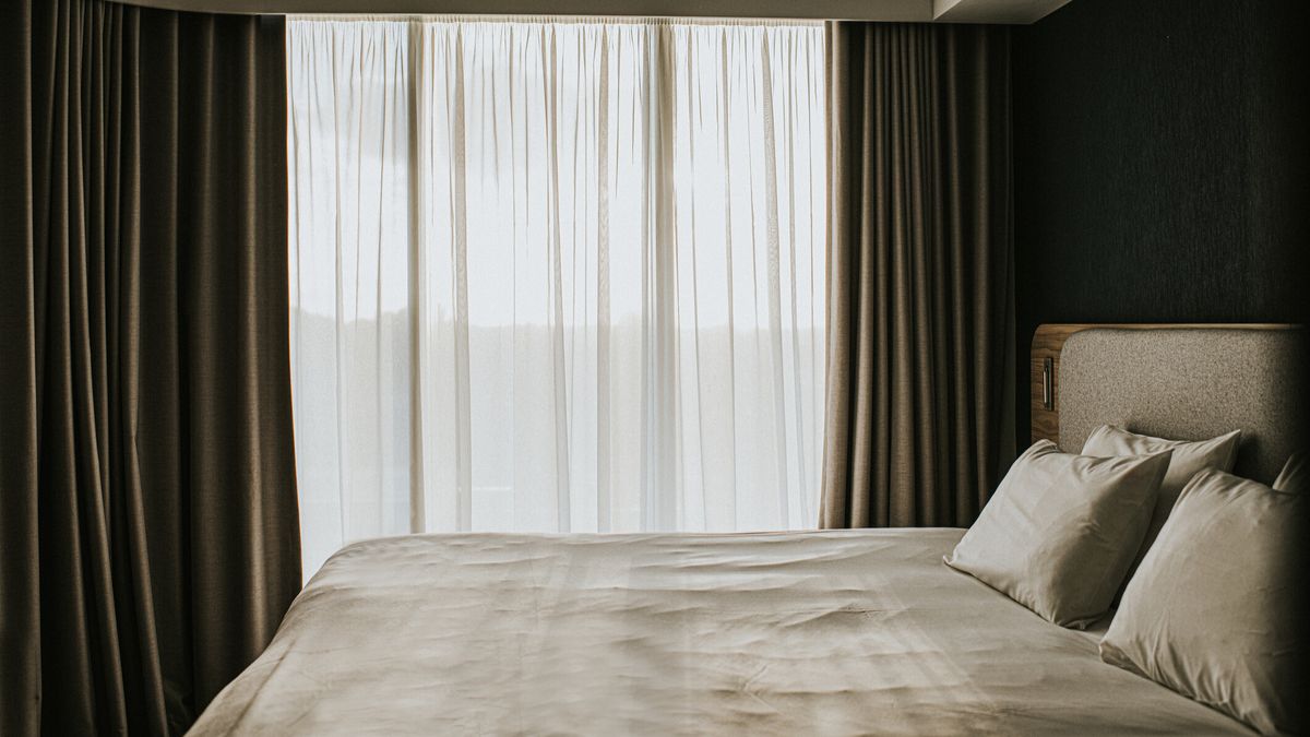 A hotel bedroom with pillows on a white bed and blinds open