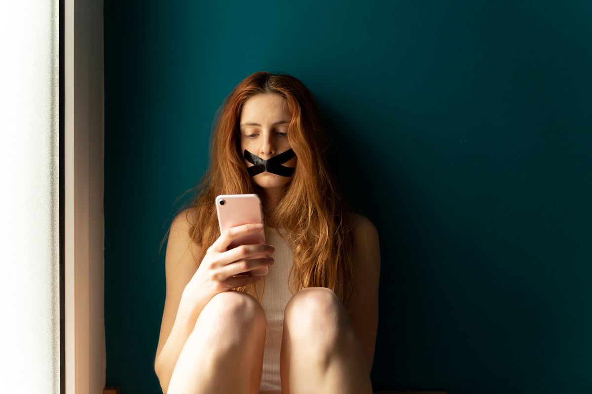 A young woman with tape on her mouth using a smartphone.