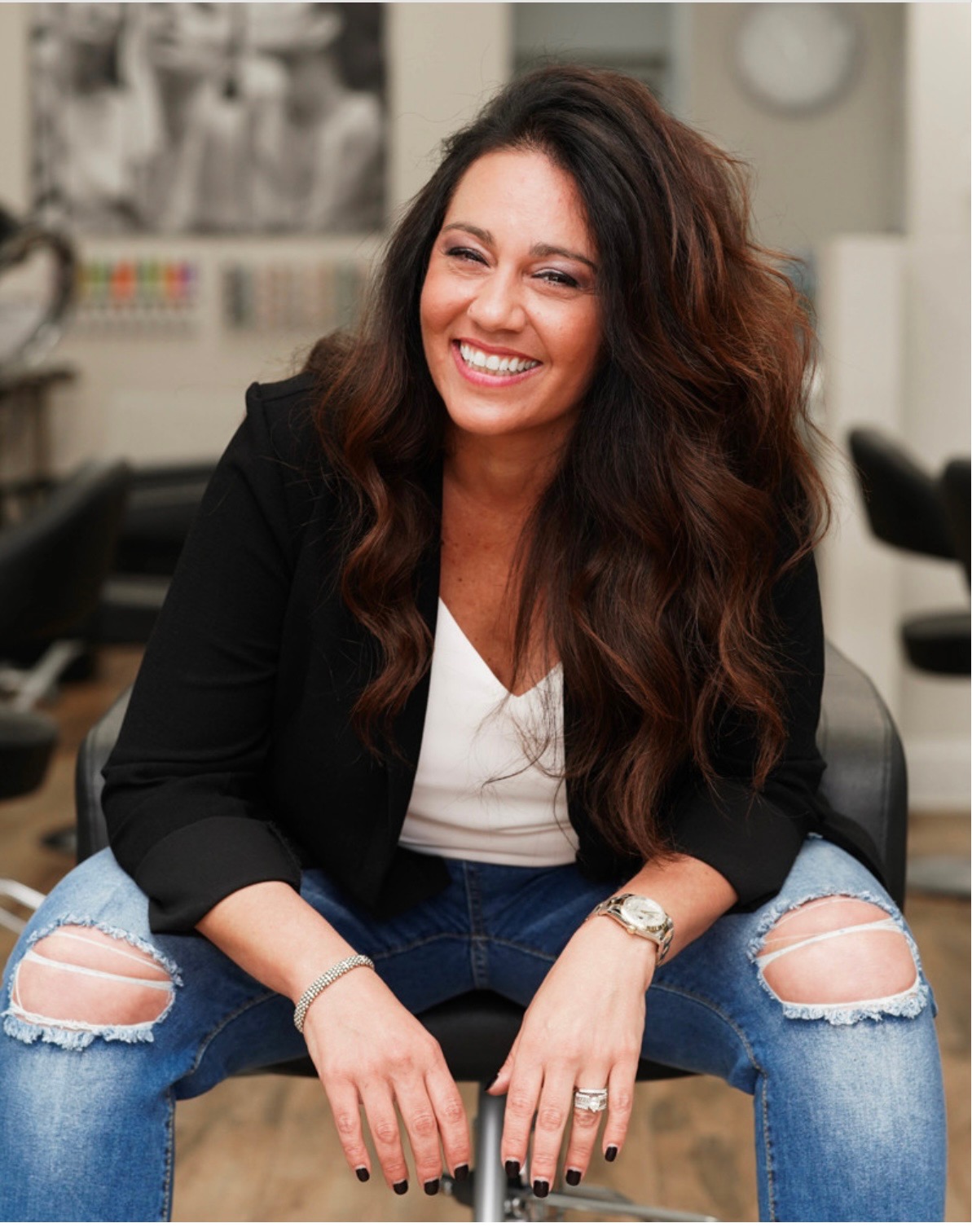 a photo of a white woman with long, brown hair wearing ripped jeans, a white top, and a black blazer. she's sitting in a chair and posing with her hands between her knees