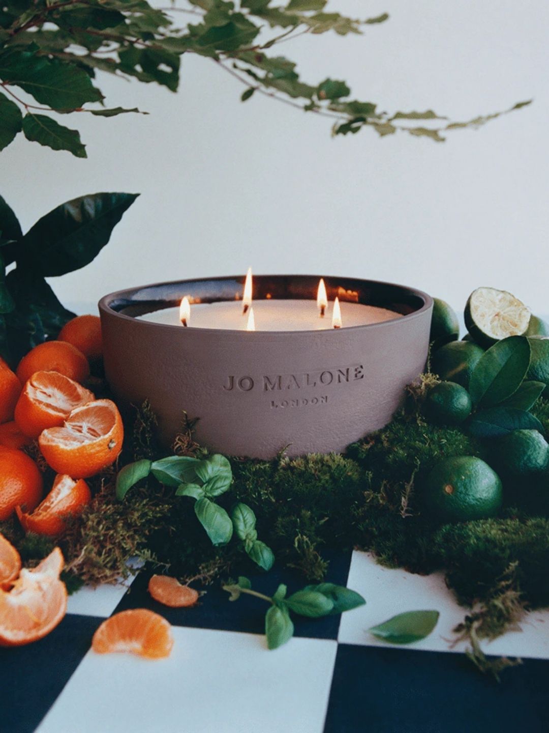 Image of a Jo Malone outdoor candle lit on a black and white checkerboard surface with oranges and limes around it.