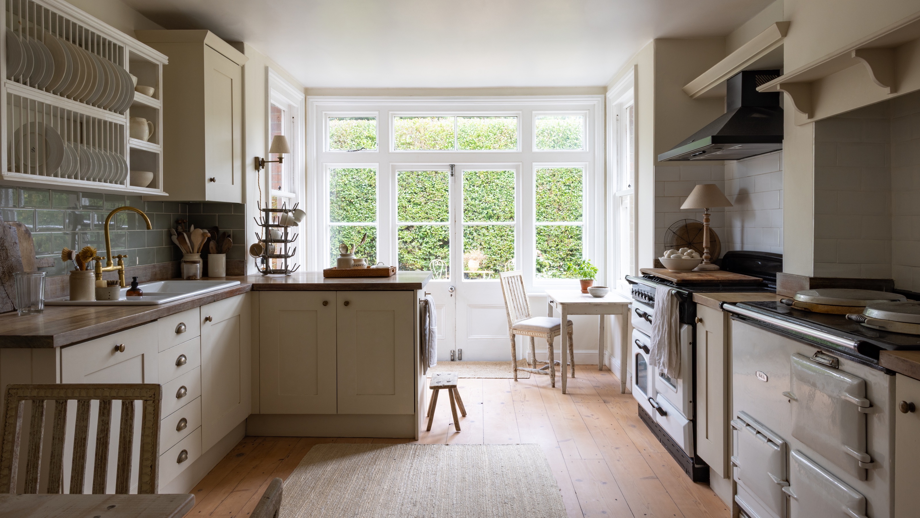 country-style kitchen with cream shaker units and range cooker, wooden floor and large windows at end of room